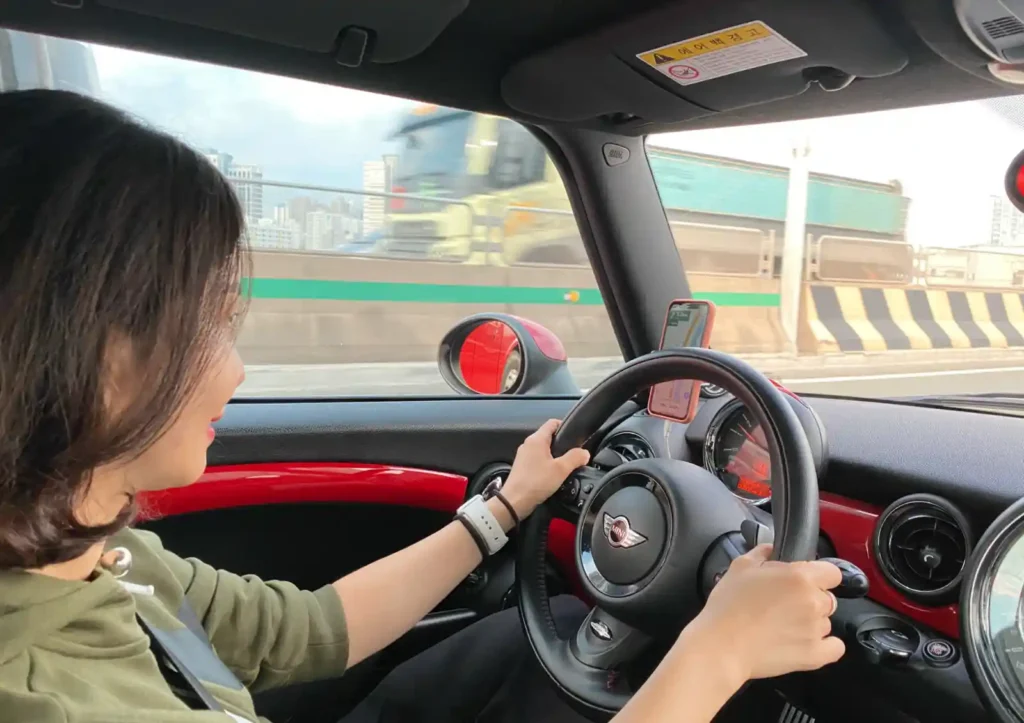 도로 위를 달리는 미니 JCW의 운전석 모습. 운전 중인 인물이 미소를 띠며 핸들을 잡고 있으며, 밝은 도심 배경과 빨간 인테리어가 어우러져 즐거운 주행의 분위기를 전한다.