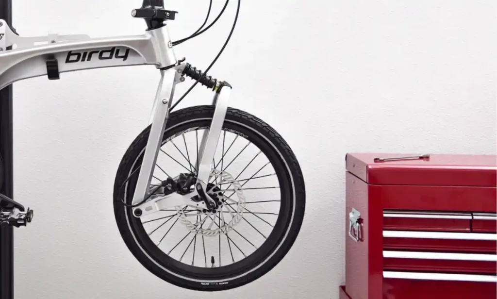 Birdy 폴딩바이크의 프론트 서스펜션과 알루미늄 프레임 구조가 클로즈업된 이미지. 스프링과 링크 구조가 뚜렷하게 보이며, 옆에는 빨간 공구함이 놓여 있다.