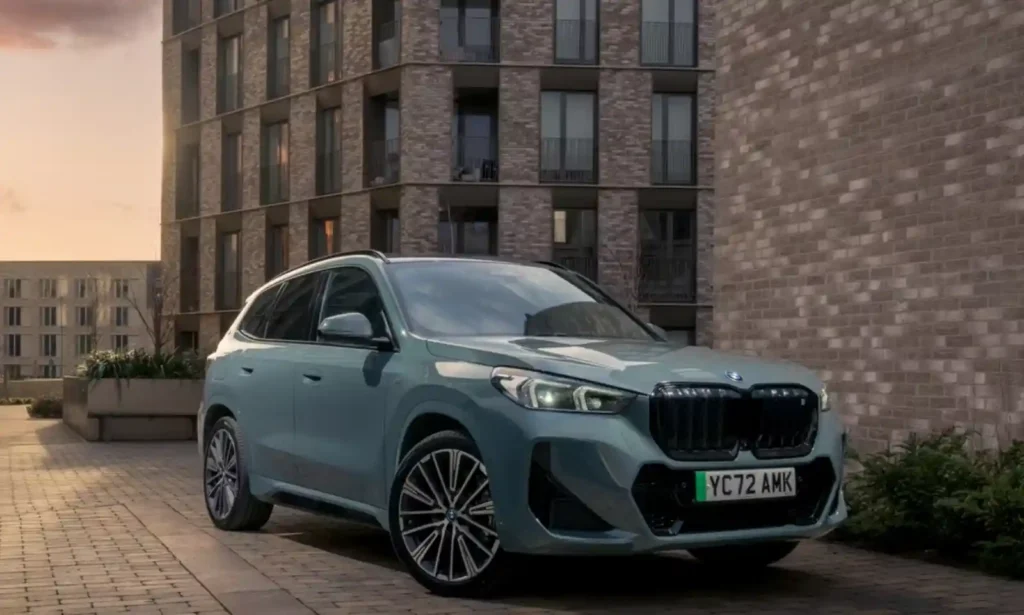 BMW iX1 전기 SUV가 도시 주거단지 앞에 주차된 모습의 정면·측면 사진으로, 컴팩트 전기 SUV의 디자인과 비율이 잘 드러난 이미지.