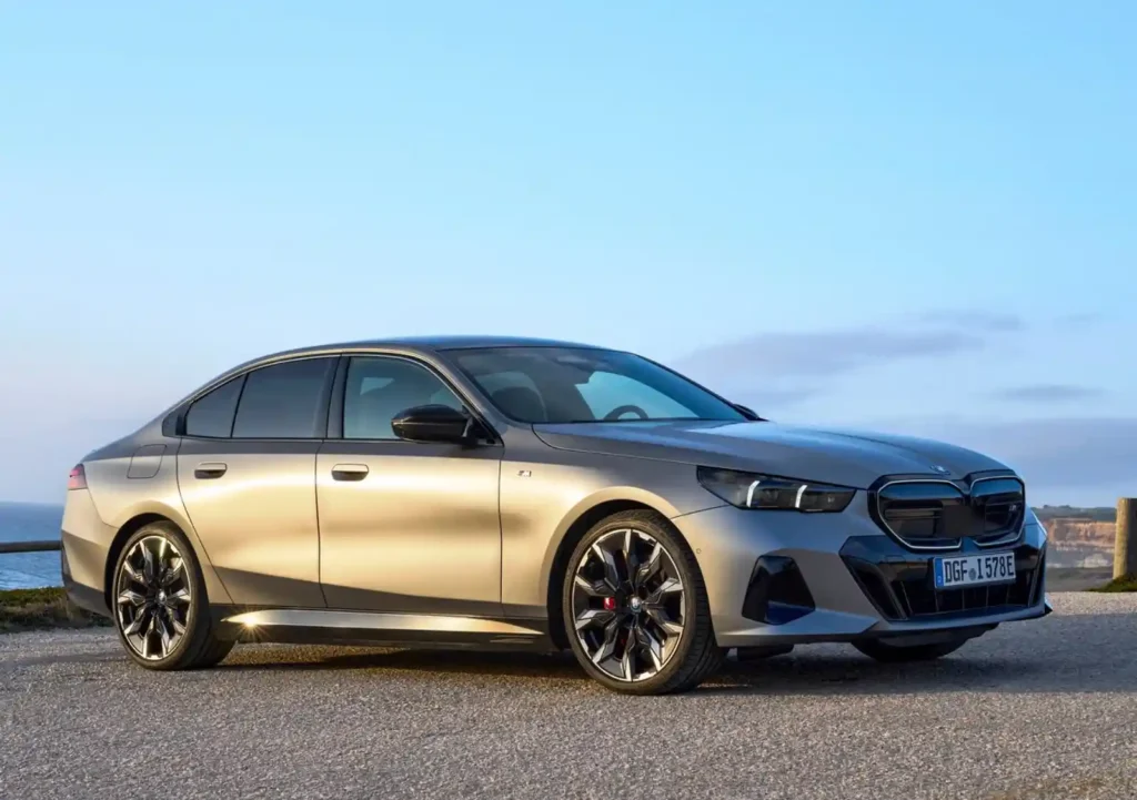 BMW i5 전기 세단이 해안 도로에서 정차한 모습으로, G60 세대의 얇은 헤드램프와 전기차 전용 그릴 디자인이 강조된 측면 이미지