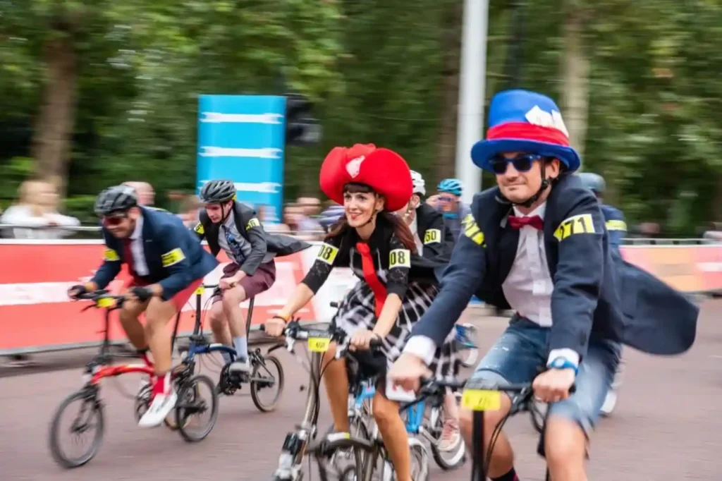 브롬톤 월드 챔피언십 참가자들이 화려한 정장과 모자를 쓰고 웃으며 달리는 장면