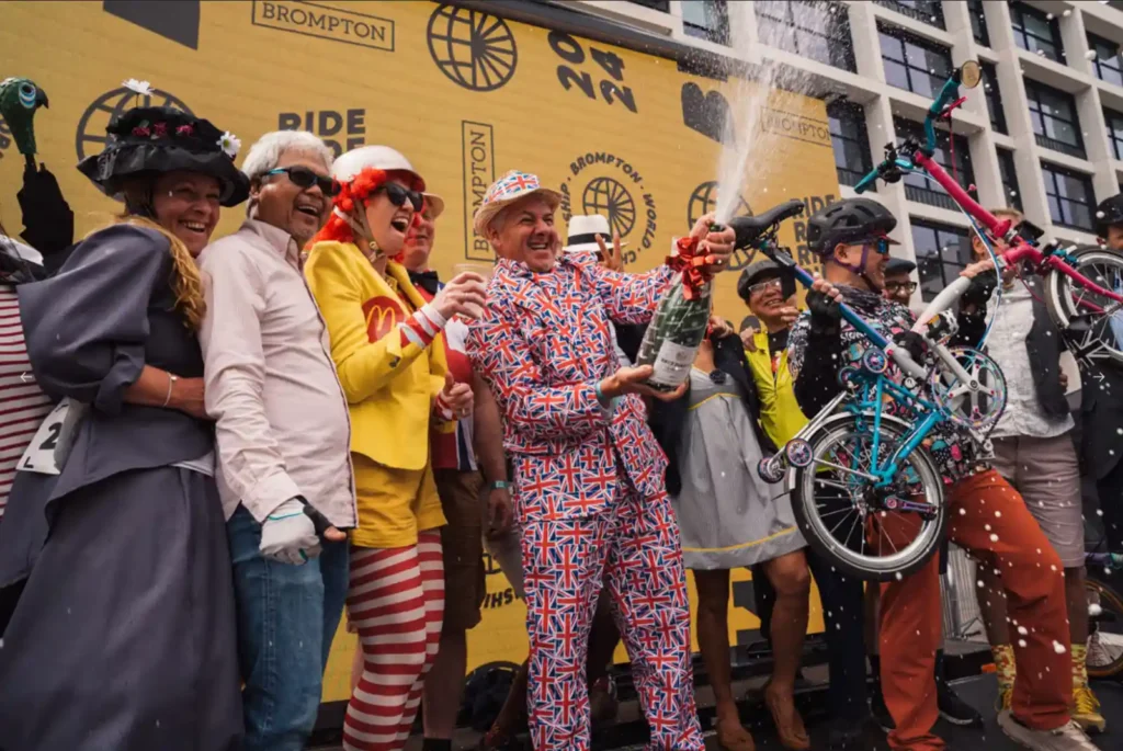 브롬톤 월드 챔피언십 참가자들이 시상식 무대에서 브롬톤 자전거를 들고 샴페인을 터뜨리며 축하하는 모습