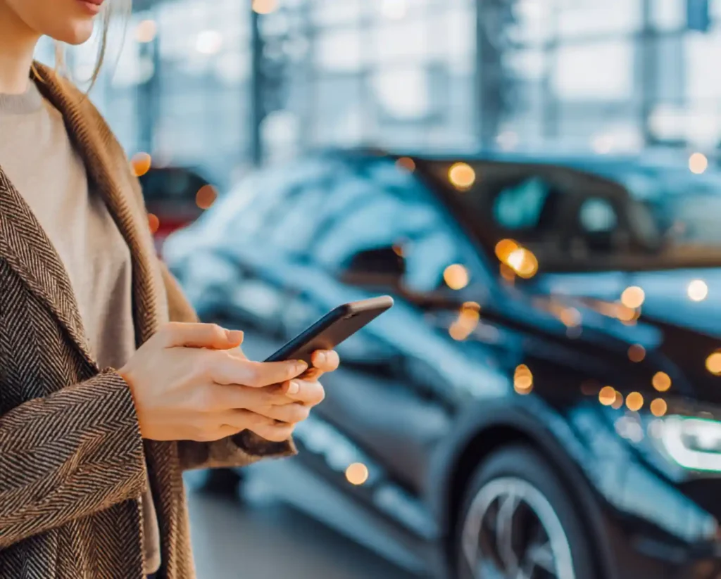 자동차 전시장 또는 정비소 앞에서 스마트폰으로 차량 수리 견적을 확인하는 사람의 모습