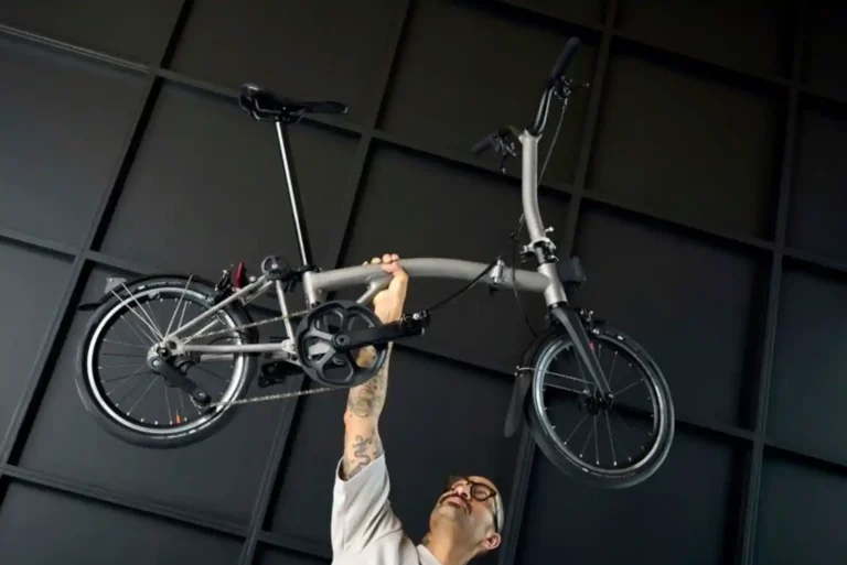 한 남성이 한 손으로 브롬톤 폴딩자전거를 들어 올리고 있습니다. 티타늄 프레임의 가벼움과 견고함을 보여주는 장면입니다.