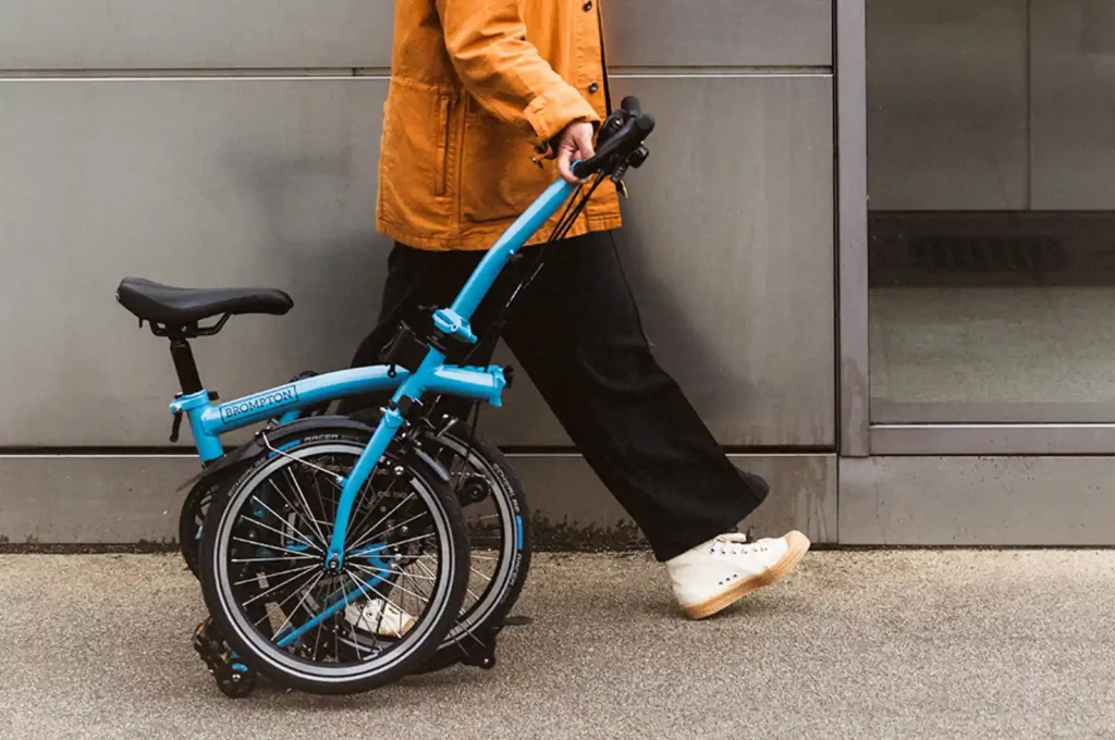 접은 브롬톤 클래식 자전거를 끌고 걷는 사람의 모습입니다. 크로몰리 스틸 프레임의 견고함과 일상 속 실용성을 보여주는 장면입니다.