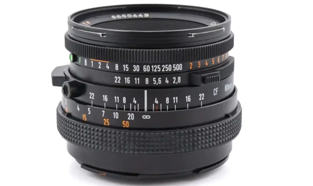 칼 자이스 플라나 80mm f/2.8 CF 렌즈가 흰색 배경 위에 놓여 있으며, 초점 거리·조리개·셔터 속도 눈금이 선명하게 보이는 클로즈업 이미지.