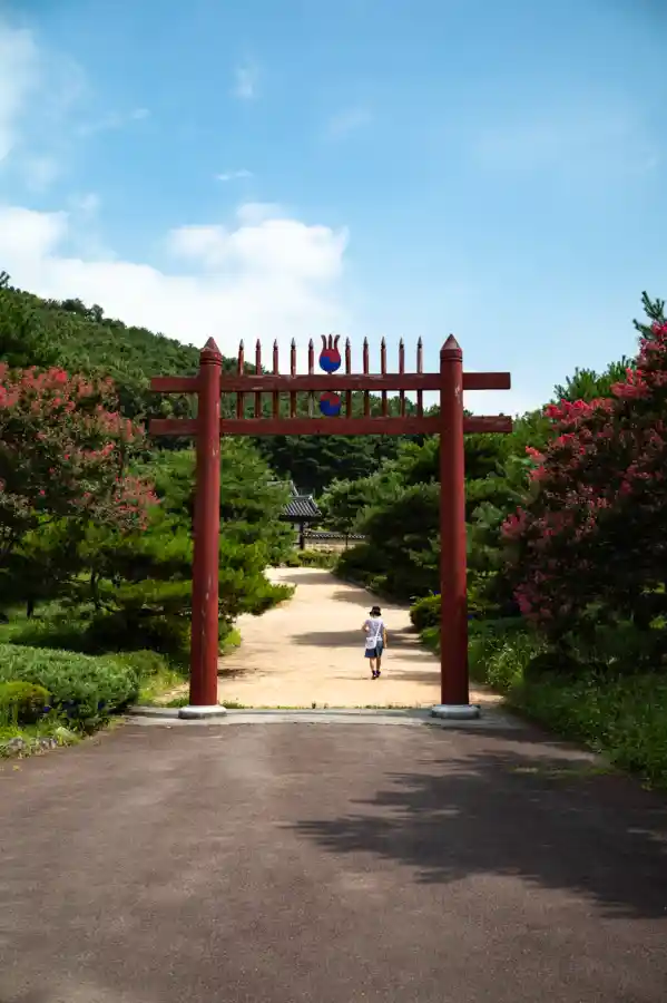 여름 하늘 아래 붉은 홍살문 너머로 이어지는 흙길을 한 사람이 걸어가는 풍경. 나무들 사이로 깊숙이 이어진 길이 고요한 분위기를 주는 야외 장면.