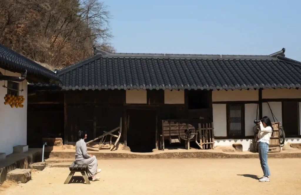 전통 한옥 마당에서 두 사람이 마주 보며 사진을 찍는 장면. 한쪽은 벤치에 앉아 있고, 다른 한쪽은 카메라를 들고 서 있는 모습. 넓은 여백과 햇살이 조용한 분위기를 만드는 Leica M10-R 촬영 사진.