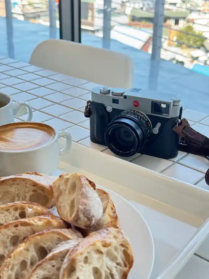 카페 테이블 위에 놓인 Leica M10-R 카메라와 커피, 그리고 바게트 조각들. 부드러운 햇살 아래 일상의 여유와 카메라의 존재감이 함께 담긴 장면.