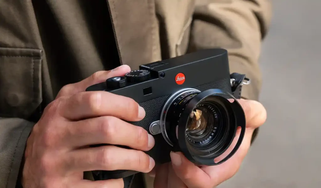 라이카 M EV1를 양손으로 쥐고 있는 모습입니다. 검은색 바디와 레드닷 로고, 장착된 Summilux 렌즈가 자연광 아래에서 선명하게 보입니다.