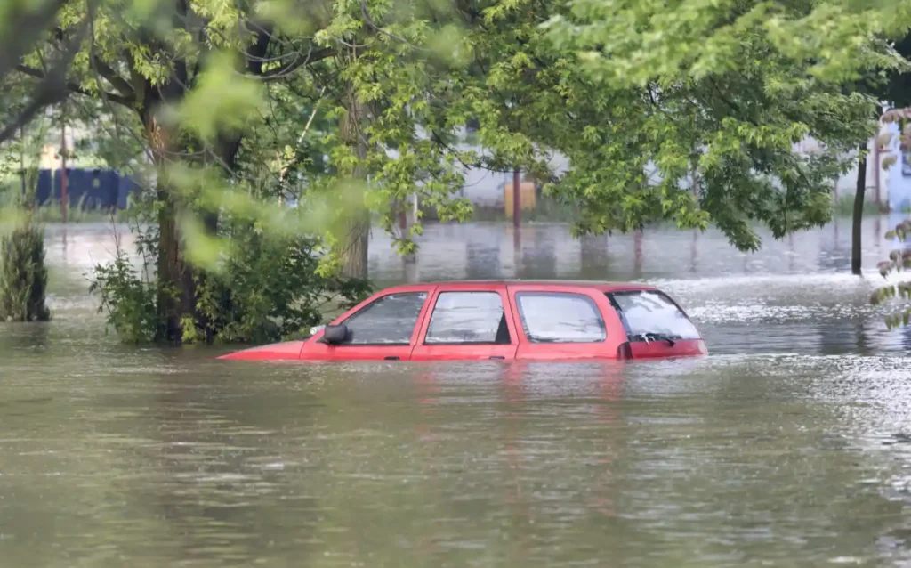 집중호우로 인한 차량 침수 피해 현장. 빨간색 SUV가 물에 거의 완전히 잠긴 채 침수된 도로에 고립되어 있으며, 주변 나무들도 물에 잠겨 있음