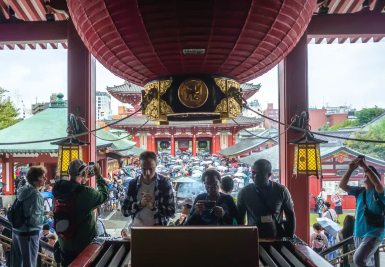 아사쿠사 센소지 호조문에서 본 본당 방향 풍경. 비 오는 날 우산 든 관광객들이 붉은 기둥과 금색 초롱 아래를 오가며, 뒤편으로 센소지 본당의 붉은 지붕과 5층탑이 보이는 모습