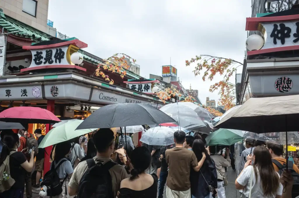 비 오는 날 아사쿠사 나카미세도리 입구 풍경. 다양한 색상의 우산을 쓴 관광객들이 좁은 거리에 빼곡히 모여 센소지 방향으로 천천히 이동하고 있으며, 양옆으로 전통 상점들과 단풍 장식이 보이는 모습