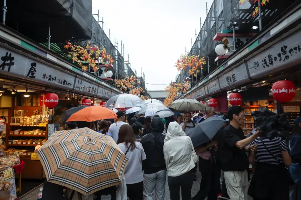 나카미세도리 내부 풍경. 양옆으로 전통 목조 상점들이 늘어서 있고, 붉은 초롱과 단풍 장식이 천장에 걸려 있으며, 비 오는 날 우산 든 관광객들이 좁은 통로를 따라 천천히 이동하는 모습