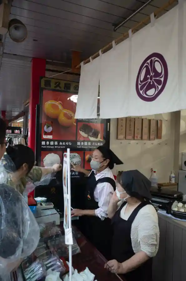나카미세도리 미타라시 당고 가게 내부. 흰색 노렌(일본 전통 천막)에 보라색 문양이 새겨져 있고, 마스크 쓴 점원들이 주방에서 당고를 준비하며 손님을 응대하는 모습