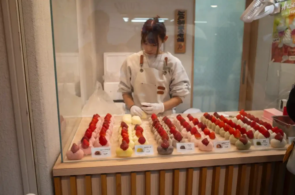나카미세도리 과일 모찌 전문점 쇼케이스. 유리 진열장 안에 딸기 모찌가 줄지어 놓여 있고, 흰색 앞치마를 입은 점원이 장갑을 낀 손으로 모찌를 정성스럽게 준비하는 모습. 핑크, 노란색, 초록색 등 다양한 색상의 모찌들이 깔끔하게 진열되어 있음