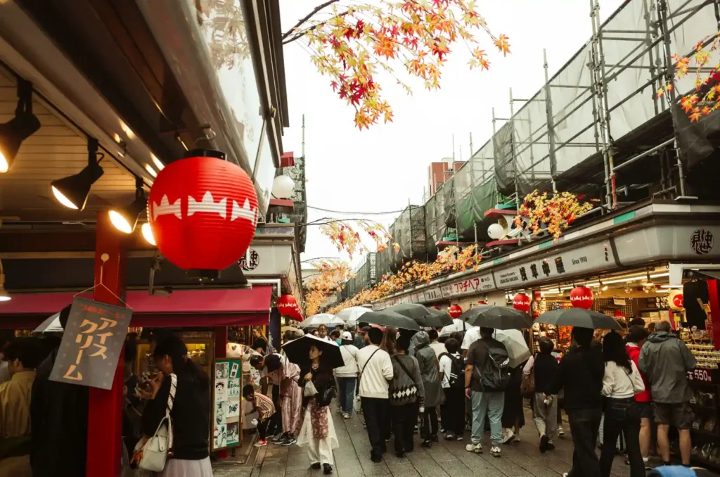 나카미세도리 거리 풍경. 붉은 초롱과 단풍 장식이 천장에 걸려 있고, 양옆으로 전통 상점들이 늘어서 있으며, 우산 든 관광객들이 좁은 통로를 따라 천천히 이동하는 모습. 회색 하늘 아래 비 오는 날의 전형적인 나카미세도리 풍경