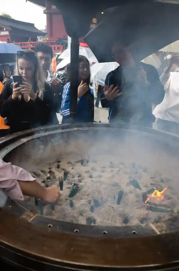 죠고로 향로 클로즈업. 거대한 청동 향로 안에서 향이 타오르며 흰 연기가 피어오르고, 사람들이 손을 뻗어 연기를 자신의 몸으로 끌어모으는 모습. 스마트폰으로 사진을 찍는 관광객들도 보임