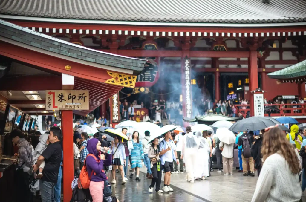 센소지 본당 앞 죠고로 향로. 거대한 향로에서 흰 연기가 피어오르고, 우산 든 수많은 관광객들이 향로 주위에 모여 연기를 쐬고 있으며, 뒤편으로 붉은 기둥의 본당 입구와 지붕이 보이는 모습