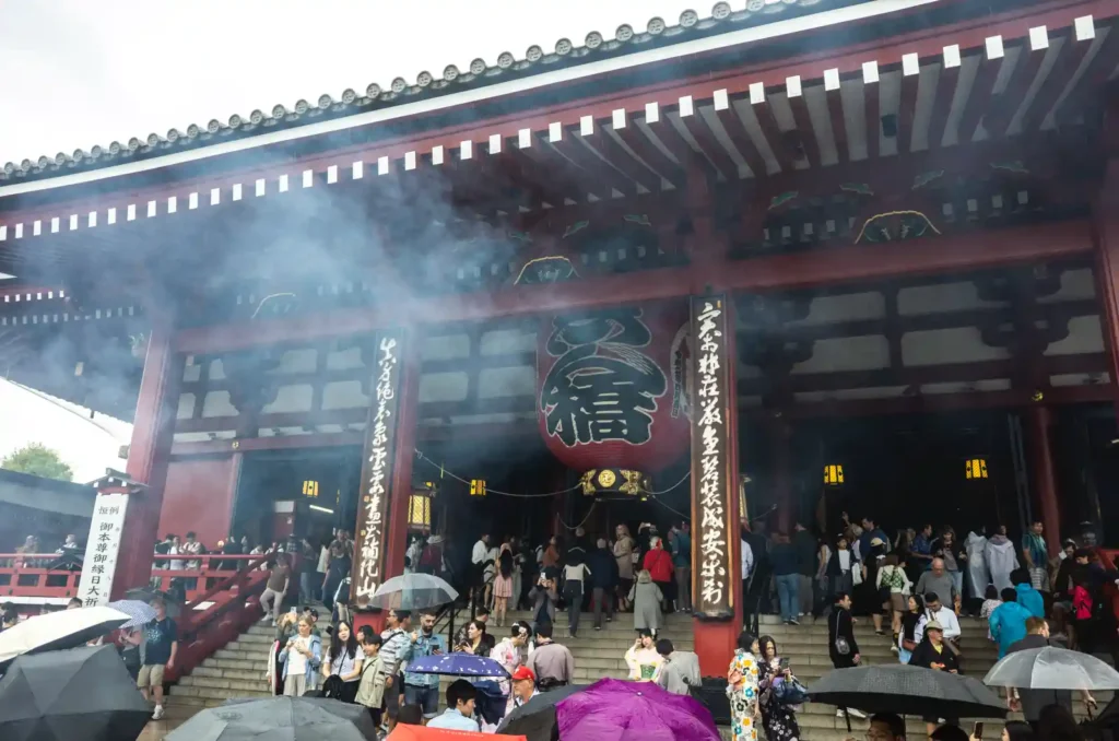센소지 본당 입구 대제등. 거대한 붉은 초롱에 '奉信' 글자가 새겨져 있고, 계단 위로 수많은 관광객들이 본당으로 들어가며, 향로에서 피어오른 흰 연기가 본당 처마 아래로 퍼지는 모습. 양옆에 한자가 새겨진 붉은 기둥이 세워져 있음