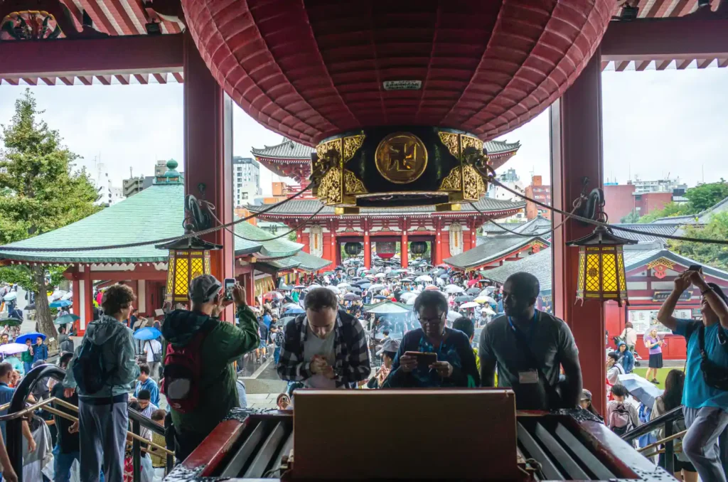 센소지 본당 내부에서 바라본 풍경. 거대한 금색 초롱이 천장에 걸려 있고, 그 너머로 호조문과 나카미세도리 방향이 보이며, 본당 안에서 참배객들이 손을 모아 기도하거나 스마트폰으로 사진을 찍는 모습. 비 오는 날 우산 든 인파가 광장을 가득 메우고 있음
