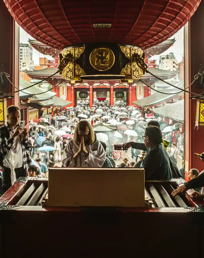 센소지 본당 내부에서 본 호조문 방향. 중앙에서 금발 여성이 두 손을 모아 진지하게 기도하고 있으며, 양옆에는 스마트폰으로 사진을 찍는 관광객들이 있고, 금색 초롱 너머로 우산 든 인파가 가득한 광장이 보이는 모습