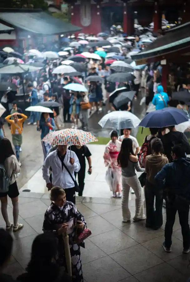 센소지 본당 처마 아래에서 본 광장 풍경. 비 오는 날 우산 든 관광객들이 광장을 가득 메우고 있으며, 전경에는 유카타를 입은 금발 여성이 스마트폰을 보고 있고, 분홍색 기모노 입은 여성과 아이들이 비를 맞으며 걷는 모습. 배경으로 호조문과 향 연기가 보임