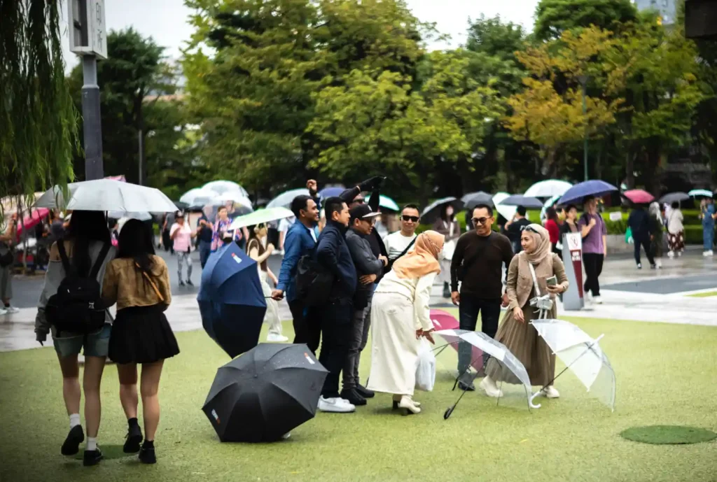 센소지 경내 잔디밭에서 사진 찍는 관광객들. 히잡을 쓴 무슬림 여성과 남성이 포즈를 취하고 있으며, 주변으로 다양한 국적의 관광객들이 우산을 쓰고 걷거나 서 있고, 배경에 녹색 나무들이 보이는 비 오는 날 풍경