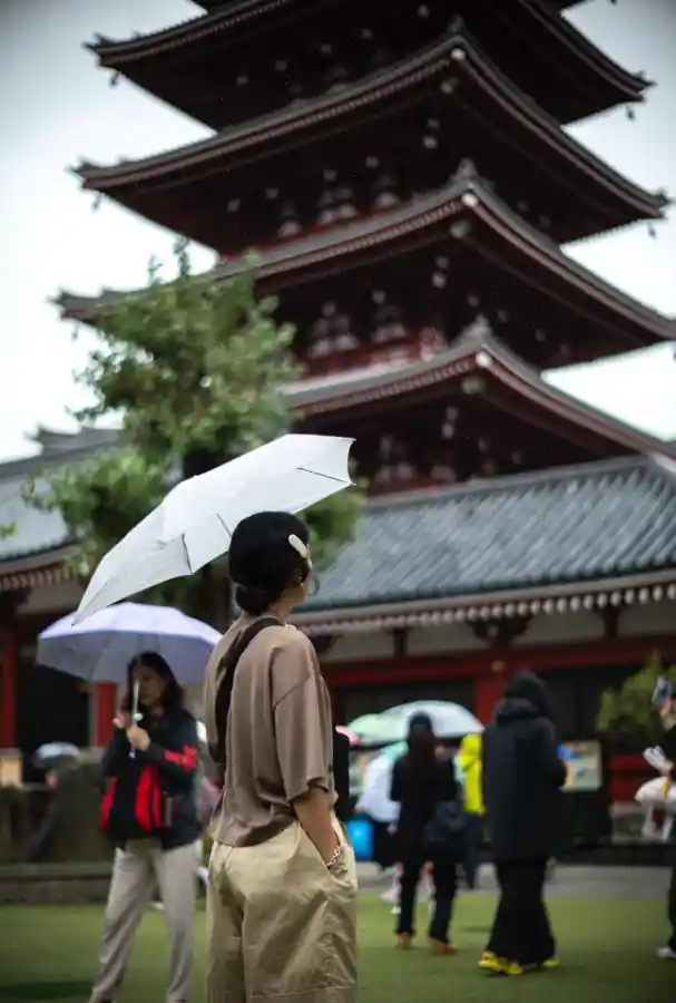 비 오는 센소지 5층탑 앞. 베이지색 옷을 입은 여성이 흰 우산을 쓰고 5층탑을 바라보고 있으며, 빗방울이 공중에 떠 있고, 배경으로 흐릿한 5층탑과 녹색 나무, 그리고 다른 관광객들의 흐린 실루엣이 보이는 모습