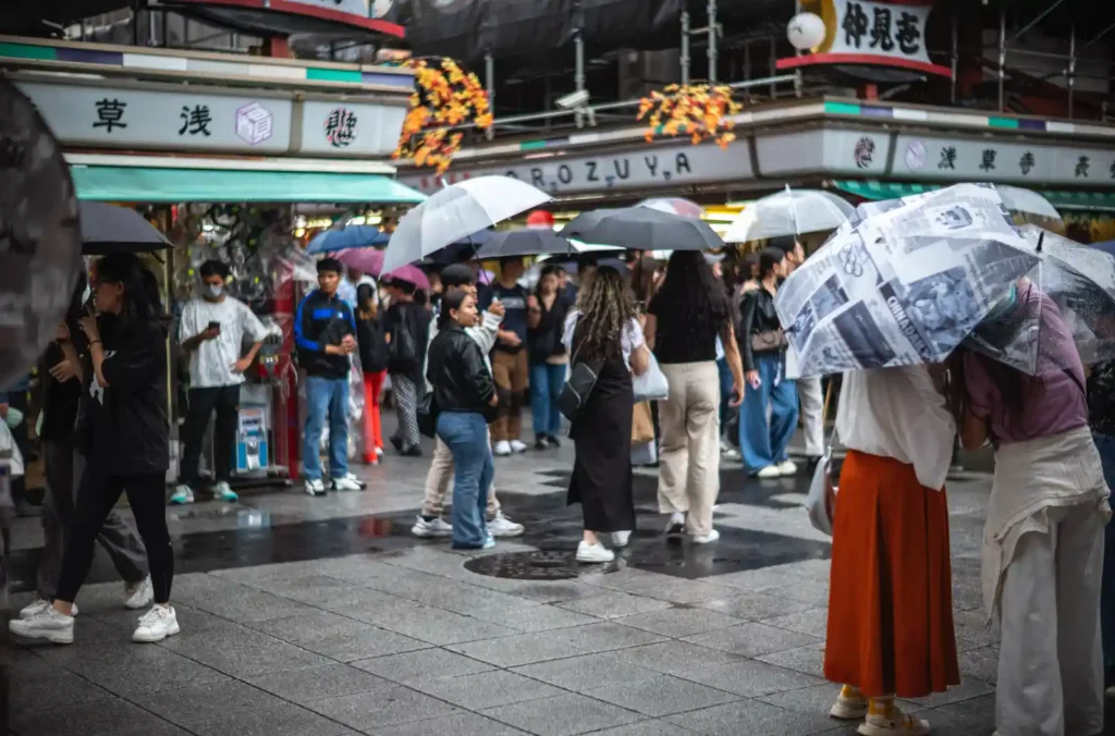 나카미세도리 귀환길. 상점 간판과 단풍 장식이 천장에 걸려 있고, 비 오는 날 우산 든 관광객들이 출구 방향으로 이동하며, 양옆으로 상점들이 영업 중인 모습. 낮 시간대 나카미세도리의 전형적인 풍경