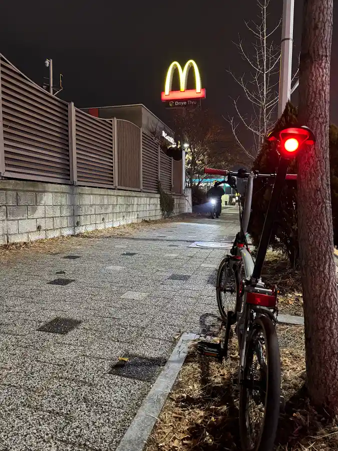 야간에 가로등 아래 나무 옆에 세워진 브롬톤 자전거, 전조등과 후미등이 켜진 상태
