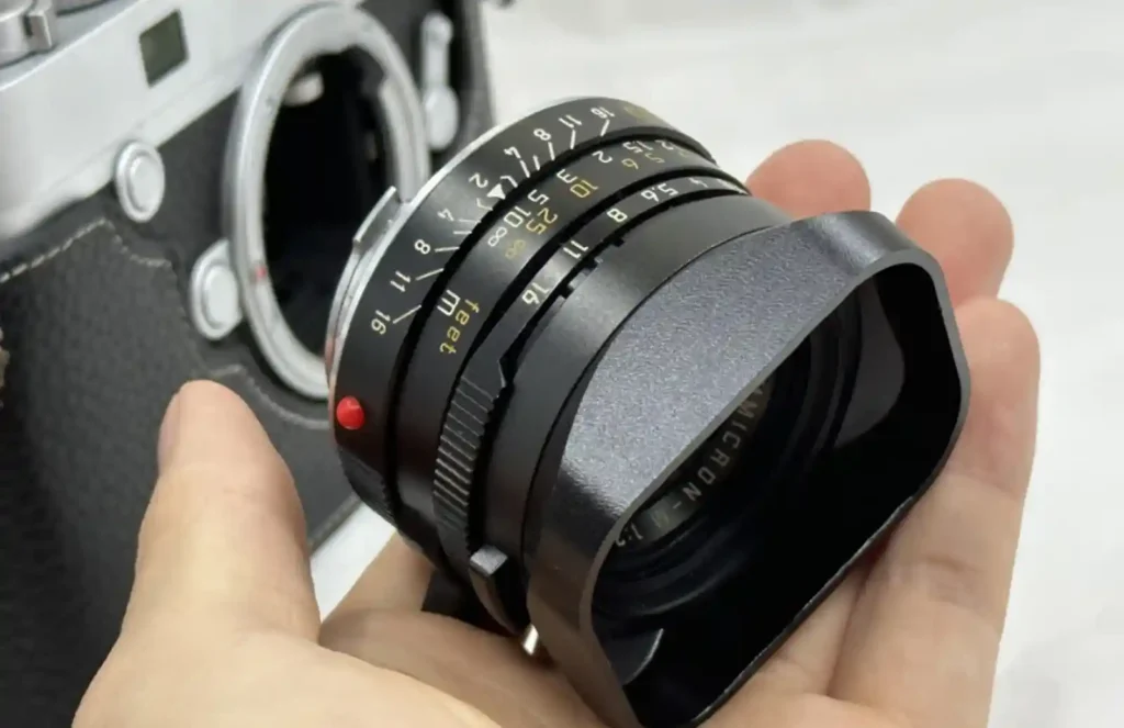 라이카 M 렌즈를 손에 올려놓고 후드 체결부를 보여주는 근접 사진으로, 금속 후드의 바요넷 구조와 정렬용 붉은 점이 선명하게 드러난 장면