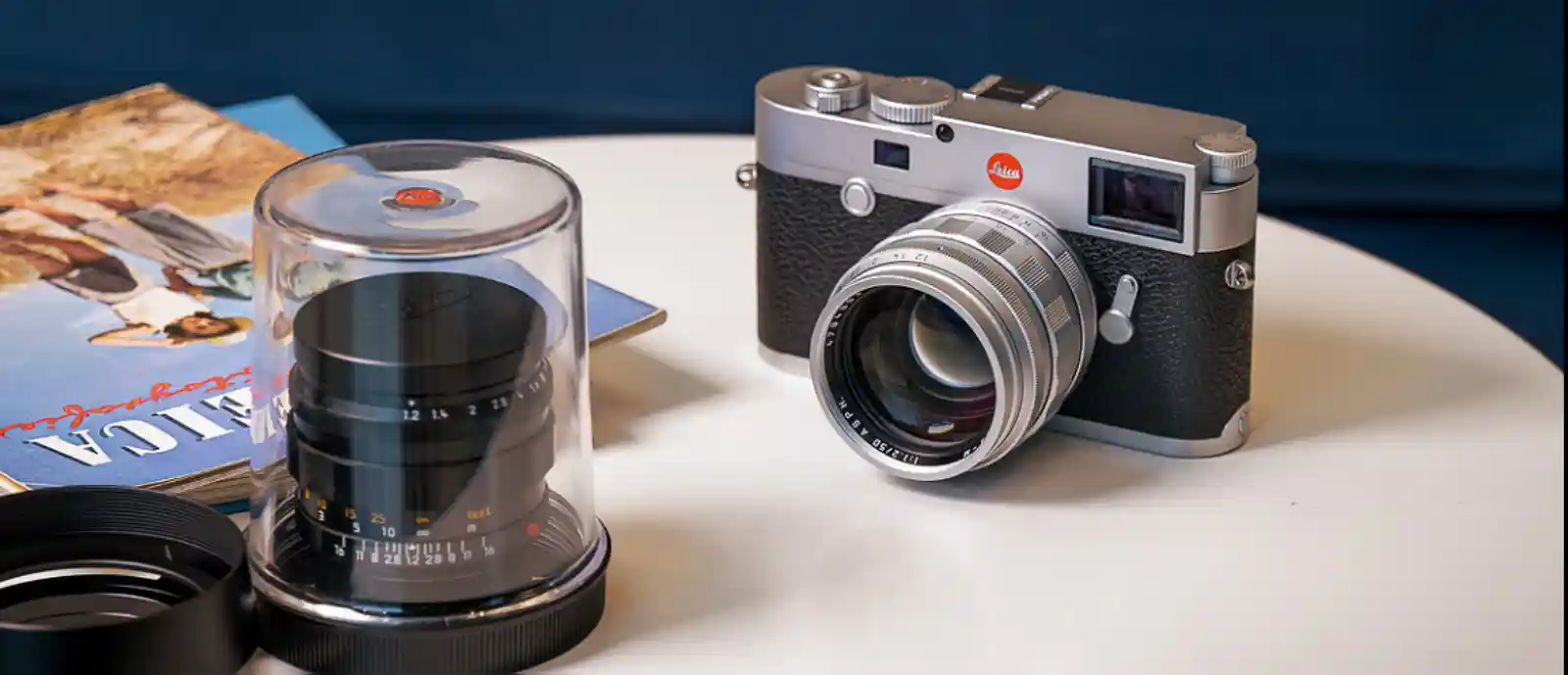 라이카 M 실버 크롬 바디에 장착된 50mm 렌즈와 투명 케이스에 보관된 녹티룩스 렌즈가 테이블 위에 놓여 있는 모습. 디지털 시대에도 작동하는 M 시스템의 호환성을 보여주는 제품 사진