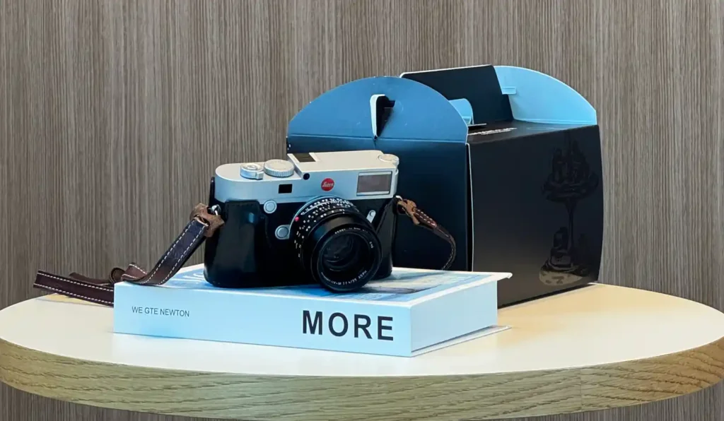 은색 상판의 라이카 M 카메라가 검은색 가죽 하프 케이스와 갈색 스트랩을 장착하고 'MORE'라고 적힌 흰색 책 위에 놓여 있다.