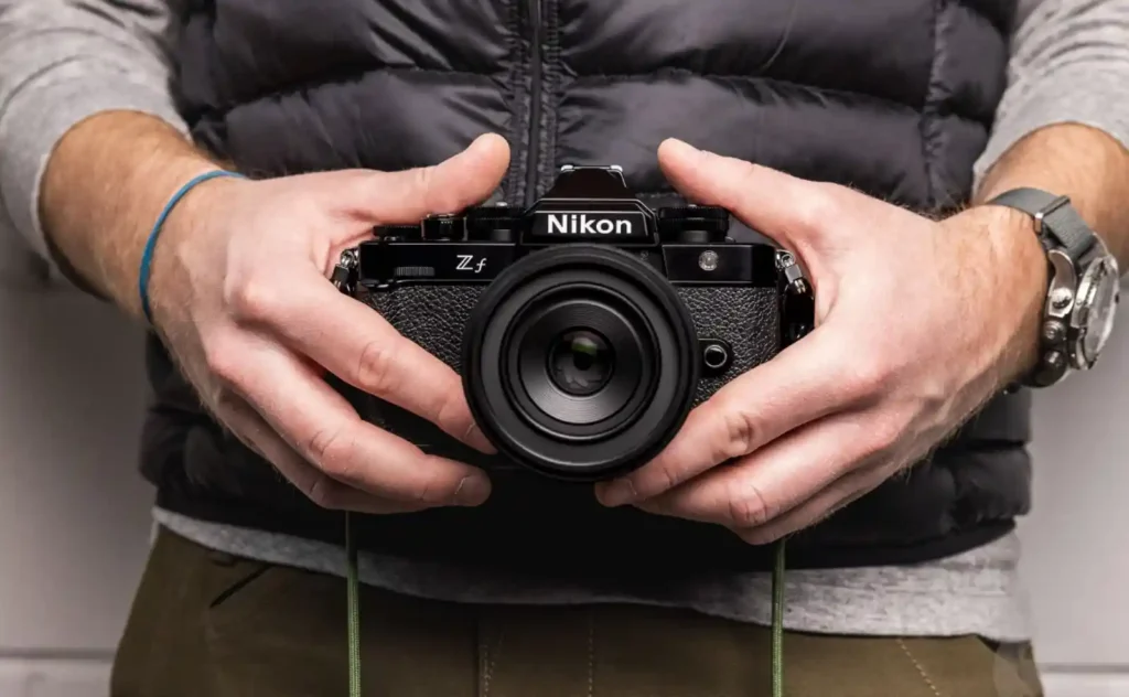 니콘 Zf 미러리스 카메라를 두 손으로 쥐고 있는 모습, 상단 황동 다이얼과 클래식한 디자인 강조
