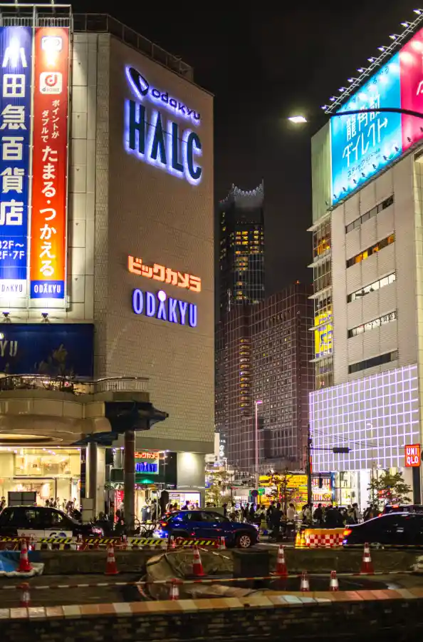 도쿄도청에서 신주쿠역으로 향하는 밤거리. 빅카메라, HALC 등 대형 건물들의 화려한 네온사인이 빛나고, 넓은 도로 위로 차량과 사람들이 오가는 신주쿠 번화가의 야경