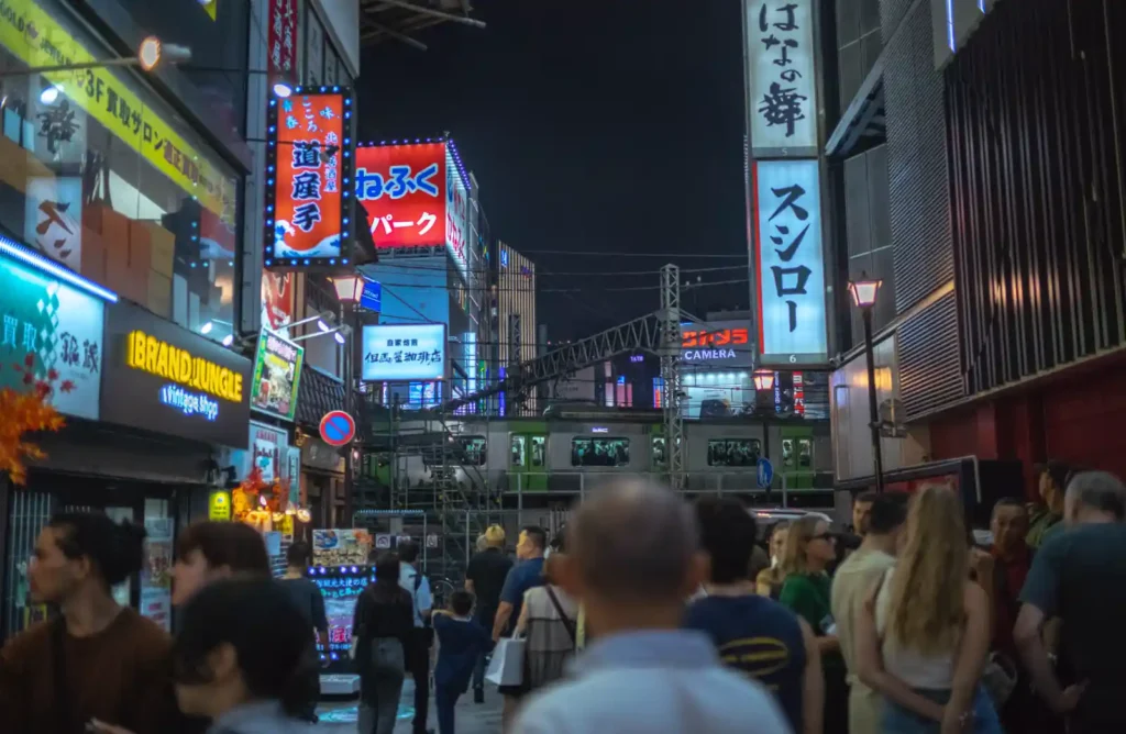 신주쿠역 서쪽 출구 인근의 밤거리. 도산테이, 스시로 등의 간판이 빛나는 좁은 상점가 거리를 수많은 관광객과 현지인들이 오가고 있으며, 뒤편으로 JR 야마노테선 전철이 지나가는 모습
