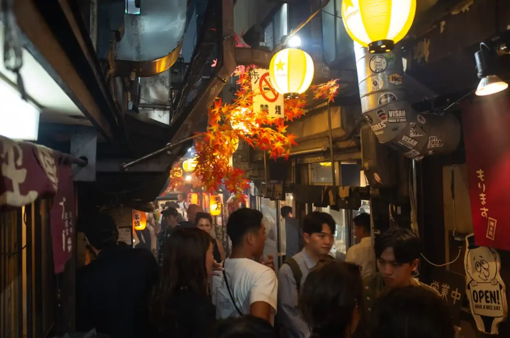 오모이데요코초 골목 내부의 밤 풍경. 노란 초롱불과 붉은 단풍 아래로 좁은 통로를 따라 수많은 사람들이 비집고 지나가며, 양옆으로 작은 이자카야 식당들이 빼곡히 들어서 있는 모습