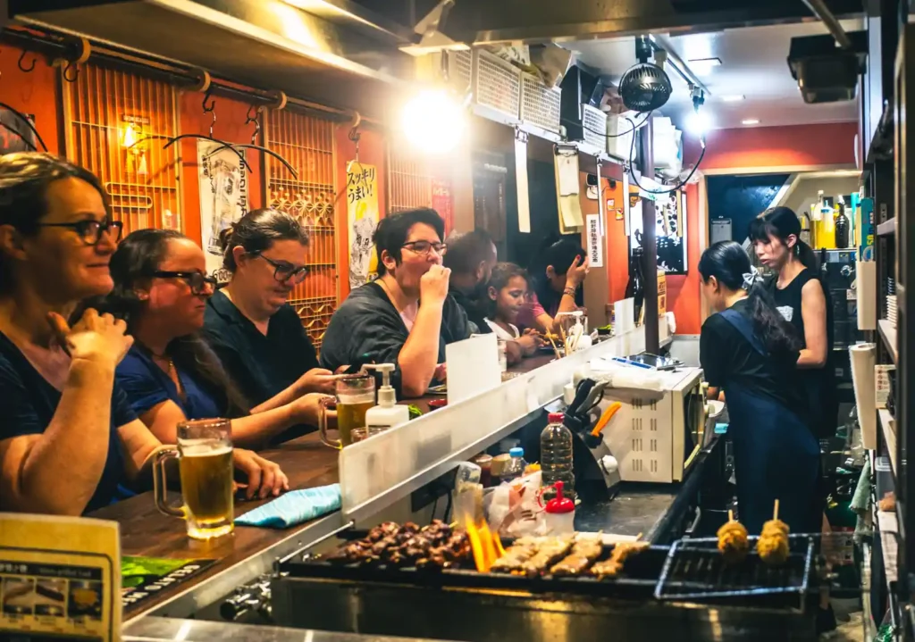 오모이데요코초 이자카야 내부. 좁은 카운터에 외국인 관광객들이 어깨를 맞대고 앉아 맥주를 마시고 있으며, 중앙 불판에서는 야키토리 꼬치가 구워지고 있고, 주인장이 요리를 준비하는 모습