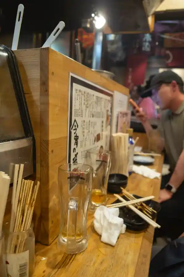 오모이데요코초 이자카야 카운터의 디테일. 나무 칸막이에 붙은 메뉴판, 젓가락통, 반쯤 마신 맥주잔, 그리고 뒤편에서 식사 중인 손님의 흐릿한 모습. 리코 GR2로 촬영한 자연스러운 일상 스냅 사진