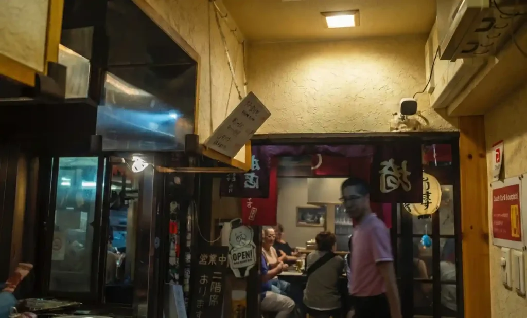 오모이데요코초 골목의 작은 이자카야 외부 모습. 노란 벽에 'OPEN' 표지판이 걸려 있고, 좁은 입구 안쪽으로 손님들이 카운터에 앉아 식사하는 모습이 보이며, 천장엔 초롱불과 메뉴판이 걸려 있는 전형적인 일본 골목 선술집 풍경