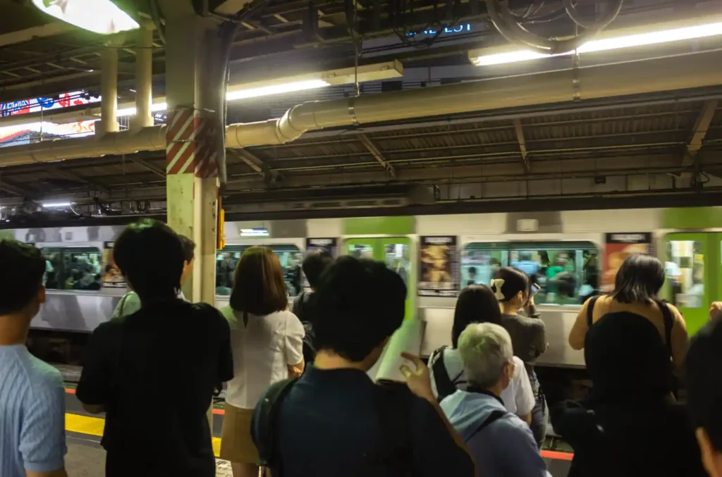 신주쿠역 지하철 플랫폼의 밤 풍경. 야마노테선 녹색 전동차가 들어오고 있으며, 플랫폼에는 수많은 승객들이 승차를 기다리며 줄지어 서 있는 모습. 늦은 시간임에도 붐비는 도쿄 지하철의 일상