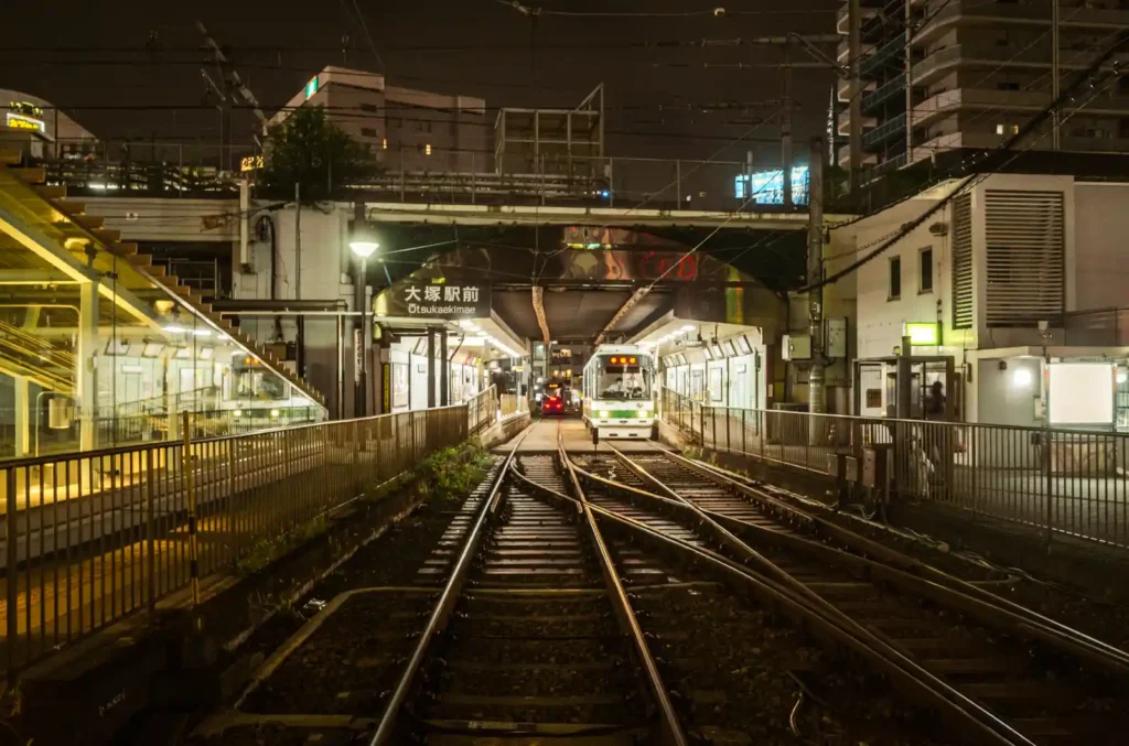 오츠카역 앞(大塚駅前 Otsukaekimae) 야경. 철길이 역 방향으로 뻗어 있고, 플랫폼에 전차가 정차해 있으며, 좌우로 주택가 건물들이 보이는 조용한 동네 역의 밤 풍경. 신주쿠의 화려함과 대비되는 평온한 오츠카의 모습