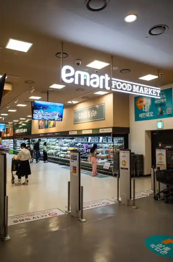 이마트 푸드마켓 수성점 매장 입구를 리코 GR2로 촬영한 사진. 천장에 emart FOOD MARKET 흰색 간판이 크게 걸려 있고, 입구 게이트를 지나면 GRAB & GO 간편식 코너와 델리 진열대가 보인다. 왼쪽에는 쇼핑카트를 끄는 고객들이 있고, 오른쪽 벽면에는 이마트 앱 광고가 붙어 있다.