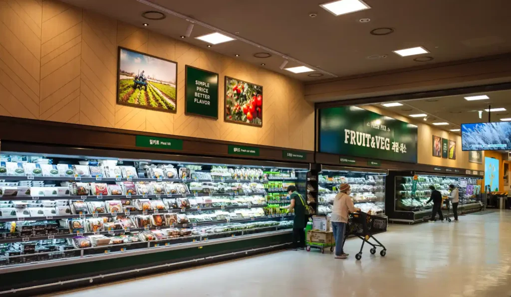 이마트 푸드마켓 채소 진열대를 리코 GR2로 촬영한 사진. 왼쪽 벽면에 SIMPLE PRICE BETTER FLAVOR 문구가 적힌 액자들이 걸려 있고, 아래 냉장 진열대에는 밀키트와 샐러드 팩들이 가지런히 정리되어 있다. 오른쪽에는 FRUIT & VEG 컬러 코스 녹색 간판이 보이고, 고객들이 쇼핑 중인 모습이 담겨 있다. 28mm 광각으로 넓은 공간감이 표현되어 있다.