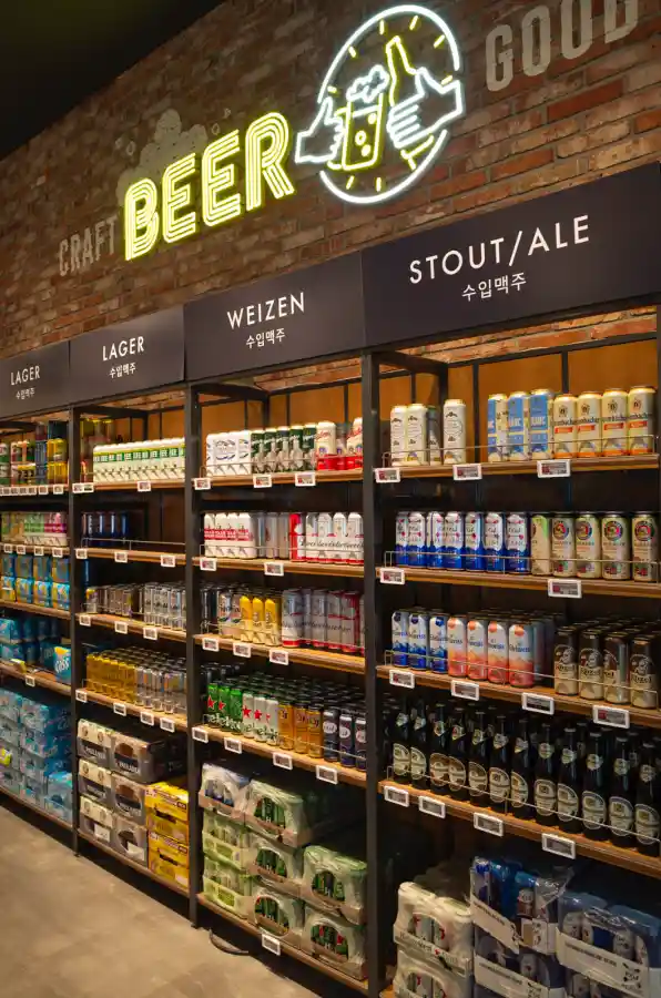 이마트 푸드마켓 맥주 코너를 리코 GR2로 촬영한 사진. 벽돌 벽면 위에 노란색 CRAFT BEER 네온사인이 빛나고, 아래 나무 선반에는 LAGER, WEIZEN, STOUT/ALE로 분류된 각국의 맥주 캔과 병들이 빼곡하게 진열되어 있다. 하이네켄, 바이엔슈테판, 파울라너 등 다양한 브랜드의 제품들이 보이며, 각 선반마다 가격표가 부착되어 있다.