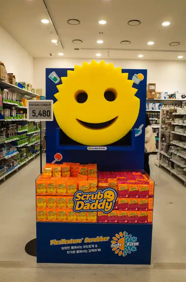 이마트 푸드마켓 생활용품 코너를 리코 GR2로 촬영한 Scrub Daddy 스크럽 스펀지 진열대. 파란색 배경에 노란색 스마일 얼굴이 크게 그려진 디스플레이가 중앙에 있고, 하단에는 오렌지색과 핑크색 Scrub Daddy 제품 박스들이 진열되어 있다. 왼쪽 위에 5,480원 가격표가 보이고, 양쪽으로 생활용품 진열대가 있다.