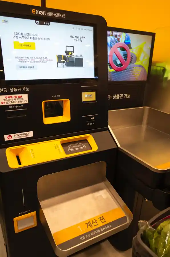 이마트 푸드마켓 셀프계산대를 리코 GR2로 촬영한 사진. 검은색 기계 상단에 바코드 스캔 시작 안내 화면이 보이고, 중앙에는 노란색 바코드 스캔 포인트가 있다. 하단에는 1. 계산 표시가 있는 바구니 놓는 공간이 보이며, 왼쪽에 CCTV 녹화 안내문, 오른쪽에 쇼핑백 사용 금지 표시가 있다. 위쪽으로 emart FOOD MARKET 로고가 보인다.