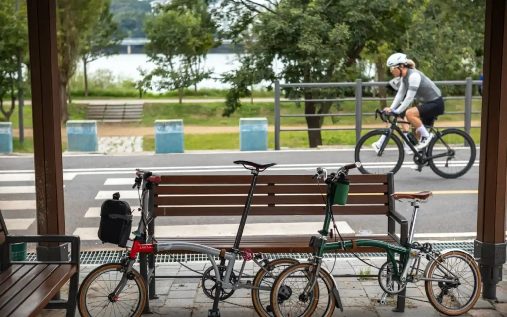 벤치에 여유롭게 주차된 브롬톤 미니벨로와 그 뒤로 빠르게 지나가는 로드바이크 라이더의 대비되는 모습