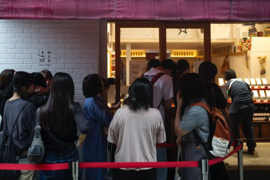 아사쿠사 우메토호시 입구에서 입장을 기다리는 손님들. 작은 식당 앞에 빨간 차단 로프가 설치되어 있고, 따뜻한 조명이 켜진 실내 안으로 여러 명의 손님들이 줄을 서 있다.